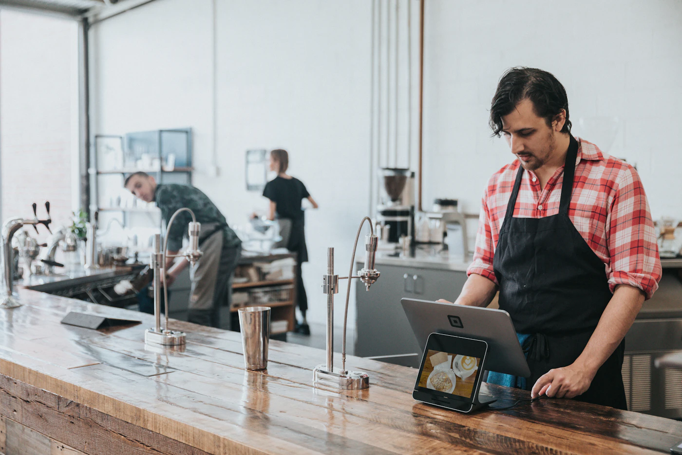 Café owner in a red check shirt and apron working on a laptop behind a wooden counter, with a card reader beside him